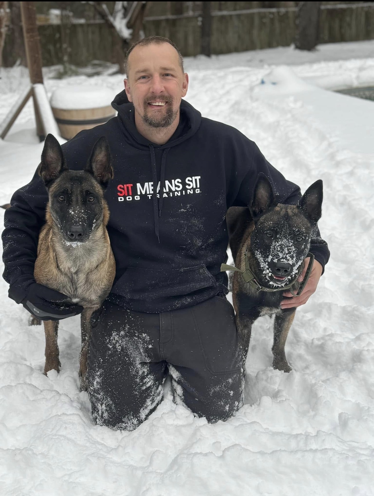 Alan kneeling in the snow with two Belgian Malinois while wearing a Sit Means Sit hoodie